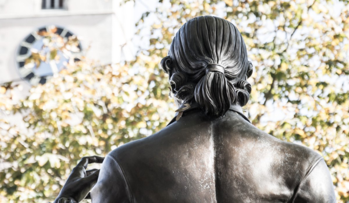 Rückansicht einer Bronzestatue mit Zopf, im Hintergrund ein Kirchturm und herbstliche Bäume., © U. Jendrass