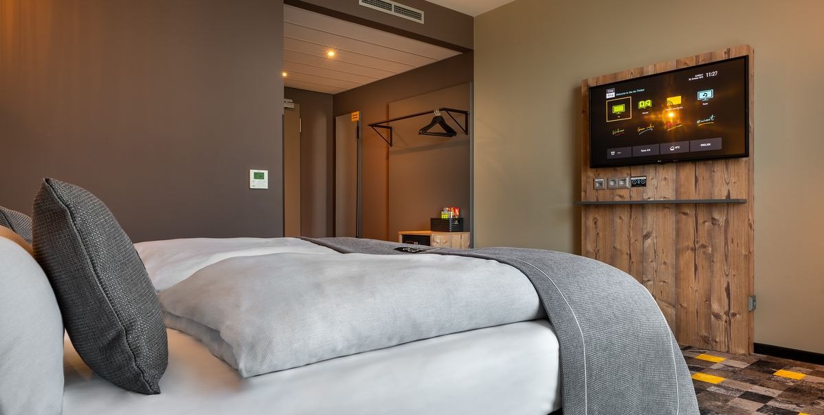 Modernes Hotelzimmer mit Doppelbett, grauen Kissen, Wand-TV und offener Garderobe. Der Boden hat ein gemustertes Design., © Novum Hospitality Modernes Hotelzimmer mit Doppelbett, grauen Kissen, Wand-TV und offener Garderobe. Der Boden hat ein gemustertes Design., © Novum Hospitality