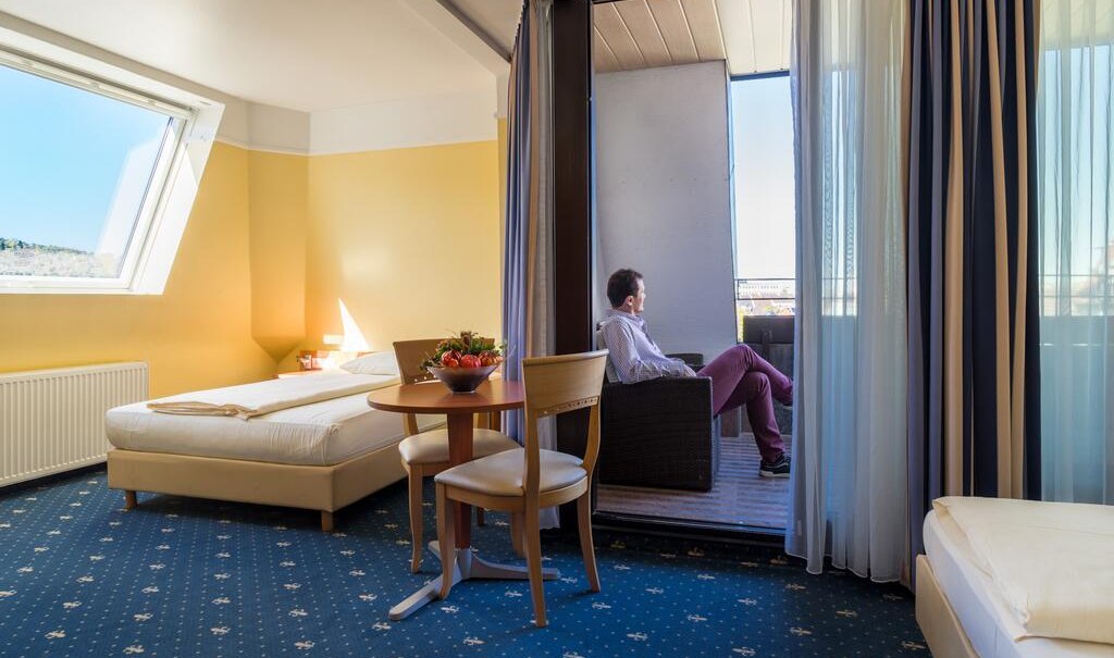 Hotelzimmer mit zwei Betten, Tisch und Stühlen. Ein Mann sitzt auf dem Balkon. Helle Wände und Teppichboden mit Muster., © Aparthotel Astro