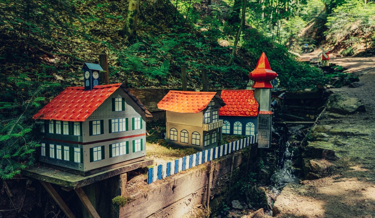 Miniaturhäuser mit roten Dächern stehen auf einer Holzplattform im Wald. Ein kleiner Bach fließt daneben. Die Szene ist bei Tageslicht aufgenommen., © Tourismus Ostalb