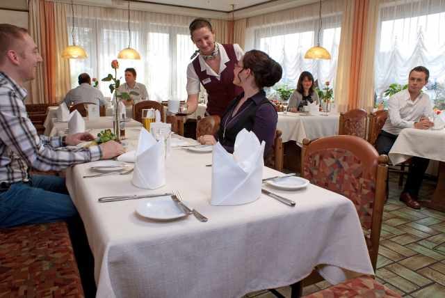 Ein Kellner bedient Gäste in einem gemütlichen Restaurant. Die Tische sind mit weißen Tischdecken und gefalteten Servietten gedeckt., © Ulli Kellenbenz Ein Kellner bedient Gäste in einem gemütlichen Restaurant. Die Tische sind mit weißen Tischdecken und gefalteten Servietten gedeckt., © Ulli Kellenbenz