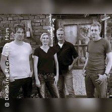 Vier Personen posieren vor einer Backsteinwand, lässig gekleidet, mit einem Fenster im Hintergrund., © links im Bild