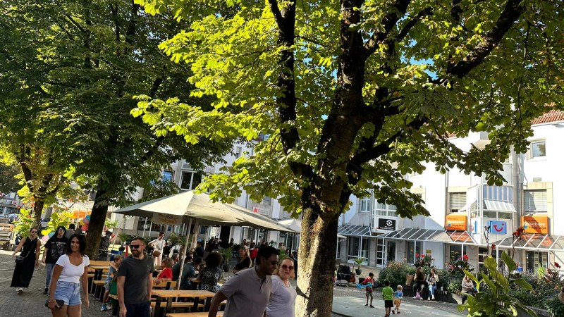 Menschen flanieren an einem sonnigen Tag durch eine belebte Einkaufsstra&szlig;e. B&auml;ume spenden Schatten, w&auml;hrend Gesch&auml;fte und Caf&eacute;s zum Verweilen einladen.