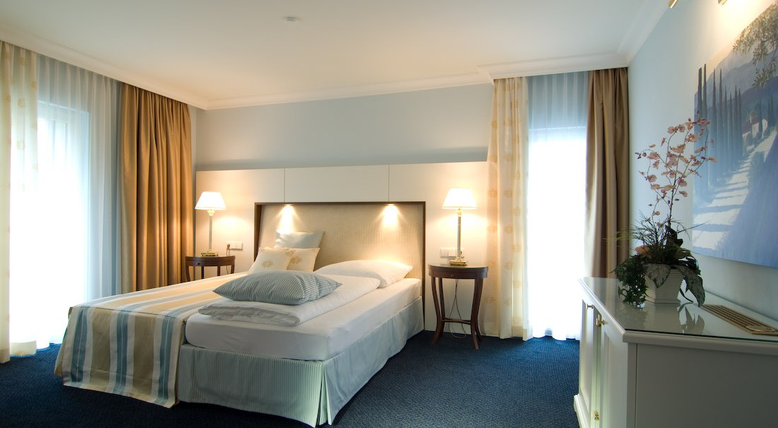 Elegantes Hotelzimmer mit Doppelbett, Nachttischen und Lampen. Helle Vorhänge und ein Bild an der Wand schaffen eine gemütliche Atmosphäre., © Jeanette Bender Elegantes Hotelzimmer mit Doppelbett, Nachttischen und Lampen. Helle Vorhänge und ein Bild an der Wand schaffen eine gemütliche Atmosphäre., © Jeanette Bender