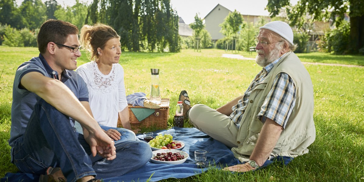 Drei Personen sitzen auf einer Decke im Park und genießen ein Picknick mit Obst und Getränken., © Jean-Claude Winkler Drei Personen sitzen auf einer Decke im Park und genießen ein Picknick mit Obst und Getränken., © Jean-Claude Winkler