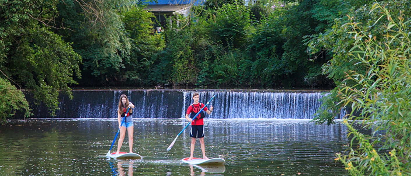 Zwei Personen paddeln auf Stand Up Paddleboards auf einem Fluss vor einem kleinen Wasserfall, umgeben von grüner Vegetation., © WTM GmbH Waiblingen