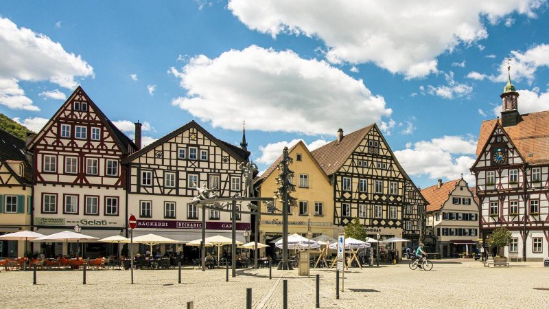 Marktplatz in Bad Urach mit malerischen Fachwerkhäusern, Cafés und Menschen bei sonnigem Wetter. Ein Radfahrer und Spaziergänger sind zu sehen., © Stuttgart-Marketing GmbH, Sarah Schmid