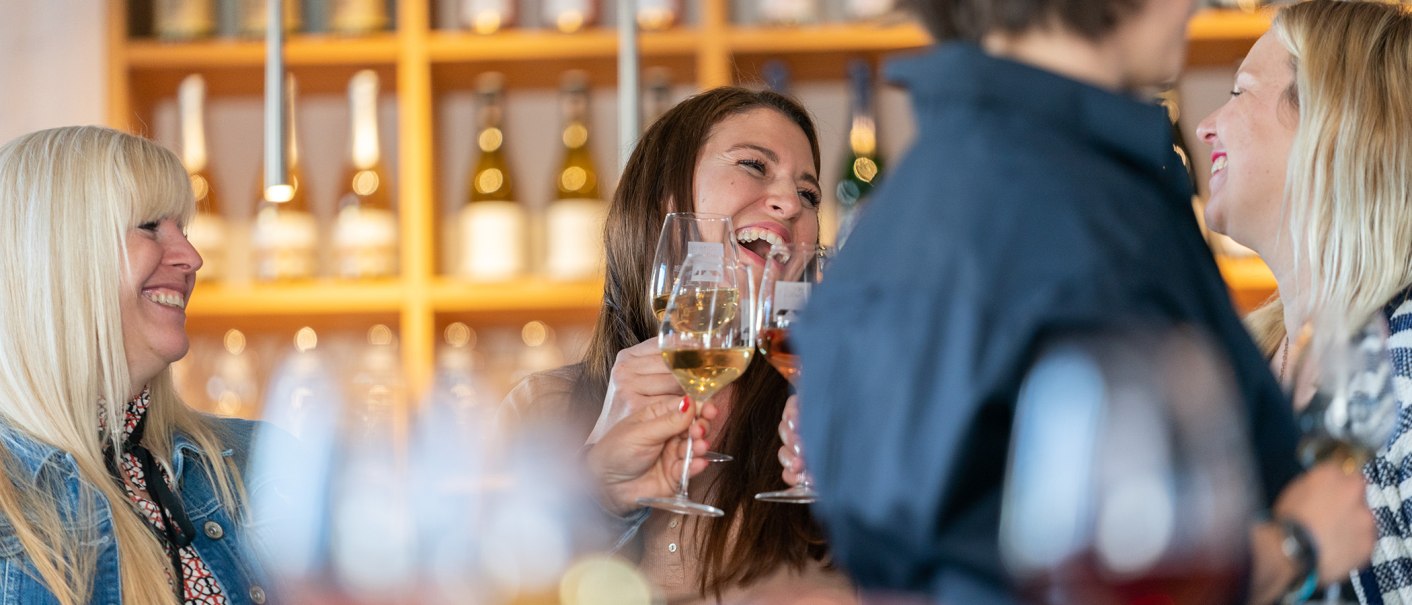Vier Personen stoßen in einer Vinothek mit Weingläsern an und lachen. Im Hintergrund sind Weinflaschen zu sehen., © Stuttgart-Marketing GmbH, Thomas Niedermüller