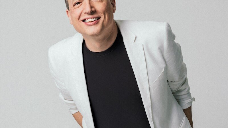 Person in weißem Anzug und schwarzem T-Shirt lächelt freundlich in die Kamera, vor einem neutralen Hintergrund., © Mauerwerk GmbH Person in weißem Anzug und schwarzem T-Shirt lächelt freundlich in die Kamera, vor einem neutralen Hintergrund., © Mauerwerk GmbH