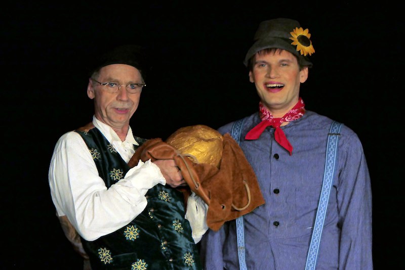 Zwei Männer in traditioneller Kleidung stehen auf einer Bühne. Einer hält einen Sack mit einem goldenen Gegenstand. Beide lächeln., © Galli Theater Backnang Zwei Männer in traditioneller Kleidung stehen auf einer Bühne. Einer hält einen Sack mit einem goldenen Gegenstand. Beide lächeln., © Galli Theater Backnang