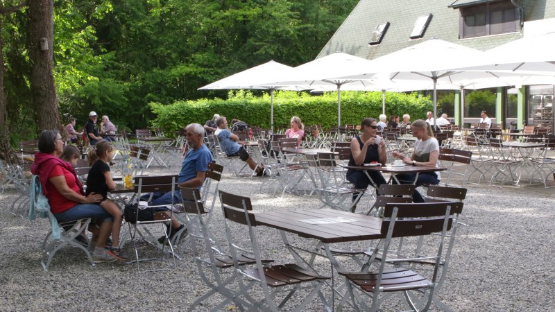 Menschen sitzen im Außenbereich eines Cafés mit weißen Sonnenschirmen, umgeben von Bäumen und grüner Vegetation., © Gabriel Holom Menschen sitzen im Außenbereich eines Cafés mit weißen Sonnenschirmen, umgeben von Bäumen und grüner Vegetation., © Gabriel Holom