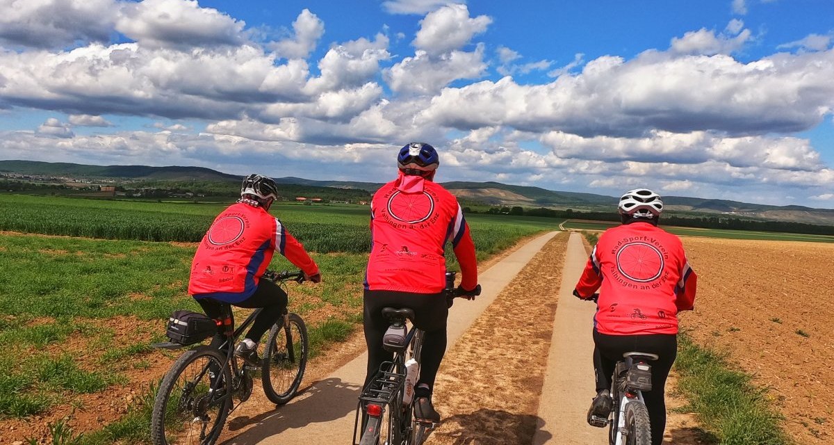 Drei Radfahrer in roten Trikots fahren auf einem schmalen Feldweg durch eine ländliche Landschaft unter blauem Himmel mit Wolken., © Land der 1000 Hügel - Kraichgau-Stromberg Drei Radfahrer in roten Trikots fahren auf einem schmalen Feldweg durch eine ländliche Landschaft unter blauem Himmel mit Wolken., © Land der 1000 Hügel - Kraichgau-Stromberg