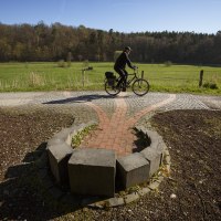 Ein Radfahrer fährt auf einem gepflasterten Weg durch eine grüne Landschaft mit Wiesen und Wäldern im Hintergrund., © VRS/ Stoppel Ein Radfahrer fährt auf einem gepflasterten Weg durch eine grüne Landschaft mit Wiesen und Wäldern im Hintergrund., © VRS/ Stoppel