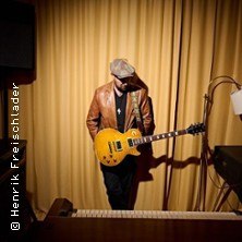 Ein Musiker mit Gitarre steht vor einem gelben Vorhang, tr&auml;gt eine braune Lederjacke und eine M&uuml;tze. Im Vordergrund ist ein Klavier zu sehen., &copy; links im Bild