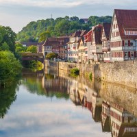 Fachwerkhäuser in Schwäbisch Hall spiegeln sich im Fluss. Eine Brücke und grüne Hügel im Hintergrund vervollständigen die malerische Szene., © Michael Kühneisen