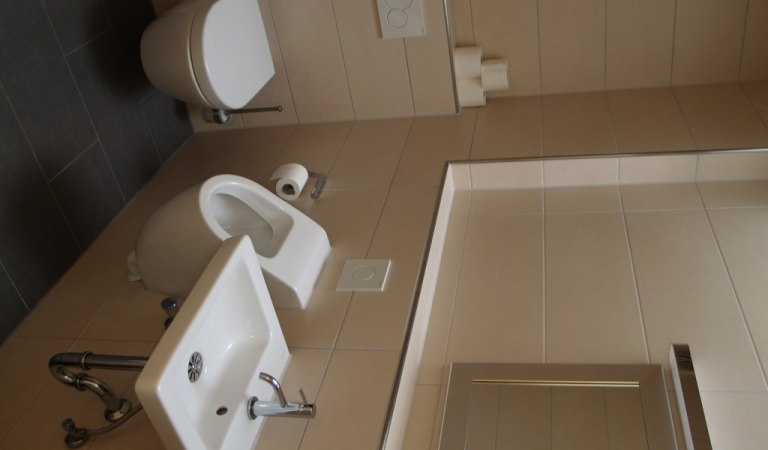 Ein modernes Badezimmer mit Toilette, Bidet und Waschbecken, alles in Weiß gehalten. Die Wände sind mit beigen Fliesen verkleidet., © TOMAS Ein modernes Badezimmer mit Toilette, Bidet und Waschbecken, alles in Weiß gehalten. Die Wände sind mit beigen Fliesen verkleidet., © TOMAS