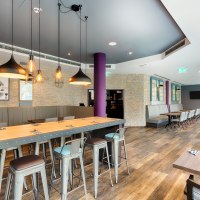 Moderner Frühstücksbereich mit langen Holztischen, Metallstühlen und stilvollen Hängelampen. Steinwände und große Fenster schaffen eine einladende Atmosphäre., © Premier Inn Germany