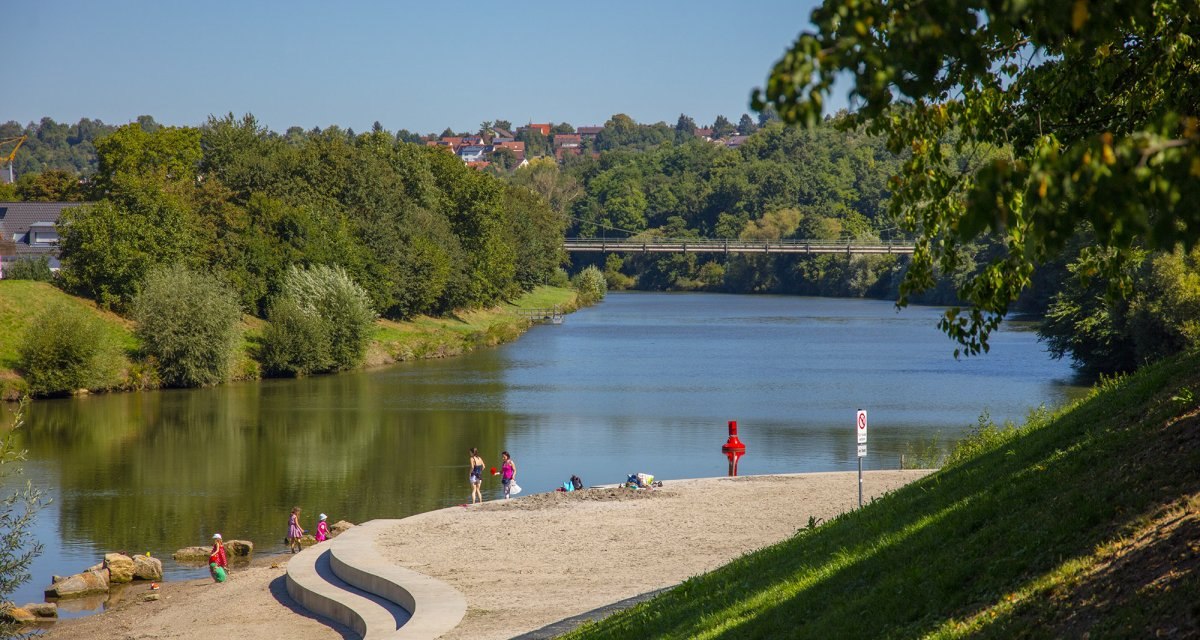 Menschen entspannen am sandigen Ufer des Remseckufers, umgeben von gr&uuml;nen B&auml;umen und einem ruhigen Fluss. Eine Br&uuml;cke ist im Hintergrund sichtbar., &copy; Remstal Tourismus e.V.