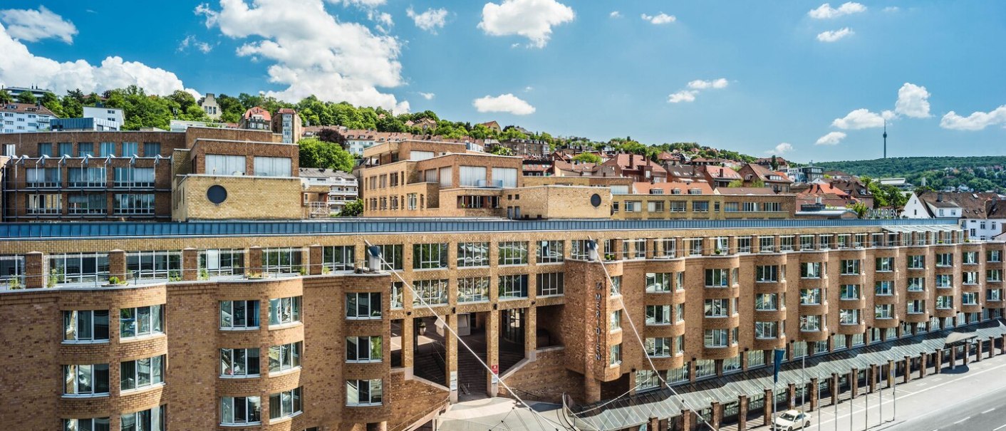 Das Le Méridien Hotel in Stuttgart mit seiner markanten Architektur, umgeben von grünen Hügeln und unter einem klaren blauen Himmel., © Hotel Le Méridien