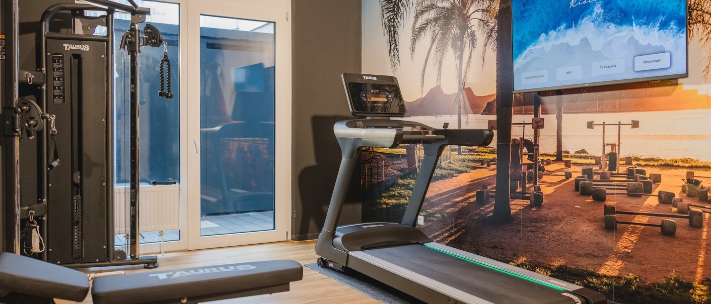 Fitnessraum mit Laufband, Kraftstation und Wandbild eines Strandes bei Sonnenuntergang. Ein Bildschirm zeigt ein Menü an. Heller Holzboden., © TOMAS Fitnessraum mit Laufband, Kraftstation und Wandbild eines Strandes bei Sonnenuntergang. Ein Bildschirm zeigt ein Menü an. Heller Holzboden., © TOMAS
