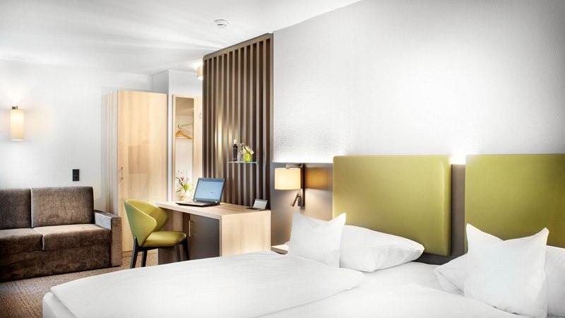Modernes Hotelzimmer mit Doppelbett, grünem Kopfteil, Schreibtisch mit Laptop, Sofa und Kleiderschrank. Helle, freundliche Einrichtung., © TOMAS
