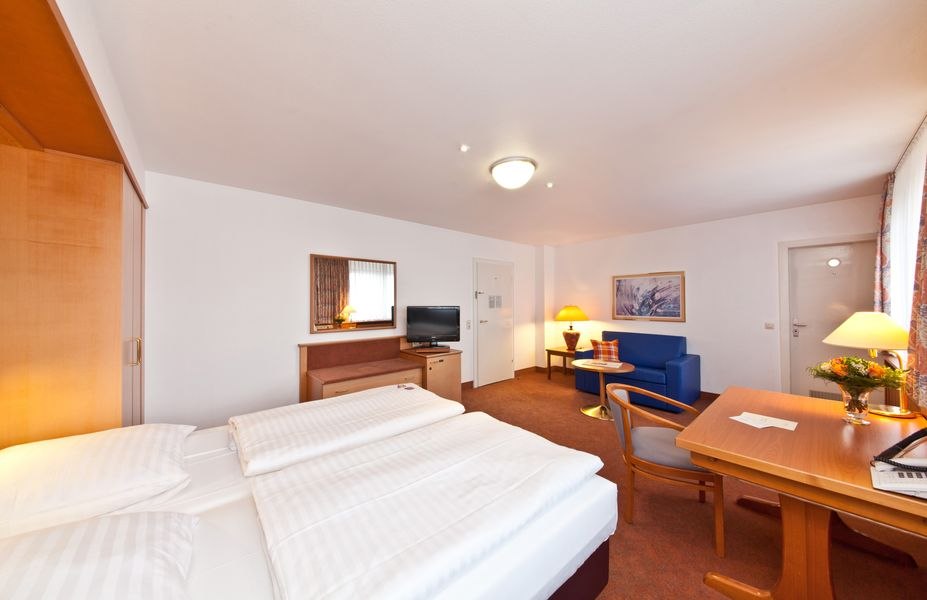 Ein geräumiges Hotelzimmer mit Doppelbett, Schreibtisch, Sofa, Fernseher und Blumen auf dem Tisch. Helle Beleuchtung und warme Farben dominieren., © Novum Boulevard Stuttgart City Ein geräumiges Hotelzimmer mit Doppelbett, Schreibtisch, Sofa, Fernseher und Blumen auf dem Tisch. Helle Beleuchtung und warme Farben dominieren., © Novum Boulevard Stuttgart City