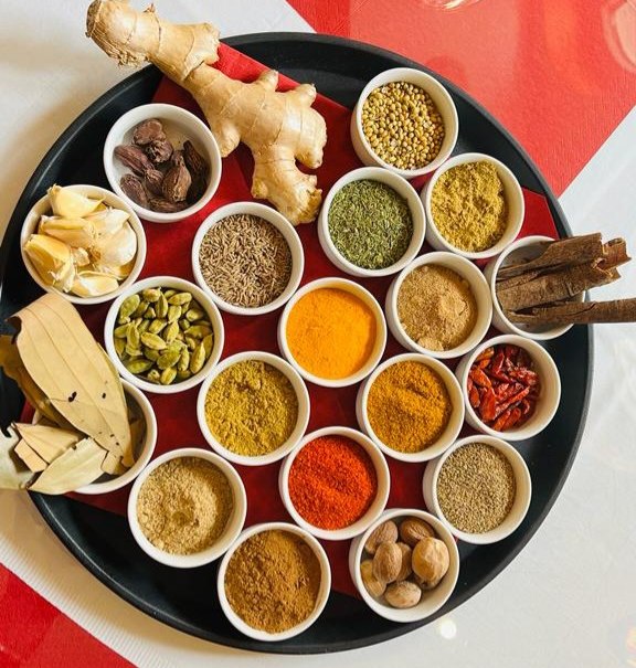 Ein rundes Tablett mit verschiedenen Gewürzen in kleinen Schalen, darunter Ingwer, Knoblauch, Zimtstangen und bunte Gewürzpulver., © Sangam