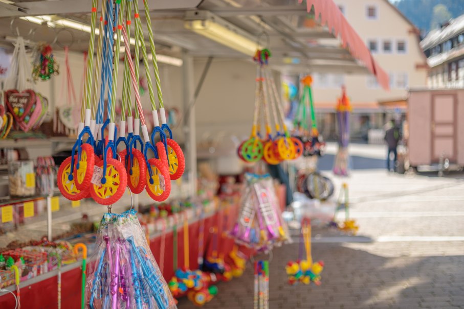 Verkaufsstand auf einem Kr&auml;mermarkt mit buntem Spielzeug und S&uuml;&szlig;igkeiten. Im Hintergrund sind Marktst&auml;nde und Geb&auml;ude zu sehen.