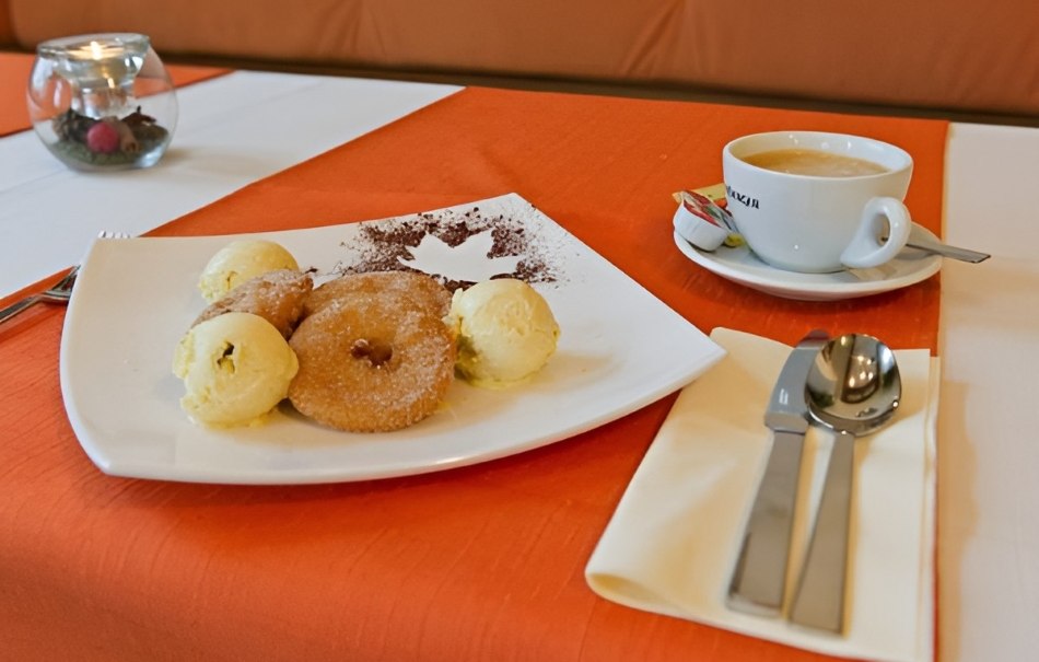 Teller mit Apfelringen und Vanilleeis, daneben eine Tasse Kaffee auf orange-weißer Tischdecke. Besteck liegt auf einer Serviette., © Hotel Neuwirtshaus Teller mit Apfelringen und Vanilleeis, daneben eine Tasse Kaffee auf orange-weißer Tischdecke. Besteck liegt auf einer Serviette., © Hotel Neuwirtshaus