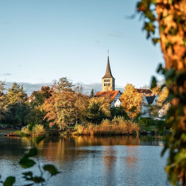 Eine malerische Kirche erhebt sich &uuml;ber herbstlich gef&auml;rbte B&auml;ume am Ufer eines ruhigen Sees, beleuchtet von warmem Sonnenlicht., &copy; Wirtschaftsf&ouml;rderung Sindelfingen GmbH