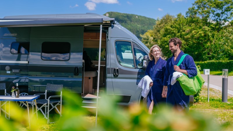 Ein Paar in blauen Bademänteln steht lächelnd vor einem modernen Wohnmobil auf einem sonnigen Campingplatz., © Thomas Niedermüller Ein Paar in blauen Bademänteln steht lächelnd vor einem modernen Wohnmobil auf einem sonnigen Campingplatz., © Thomas Niedermüller