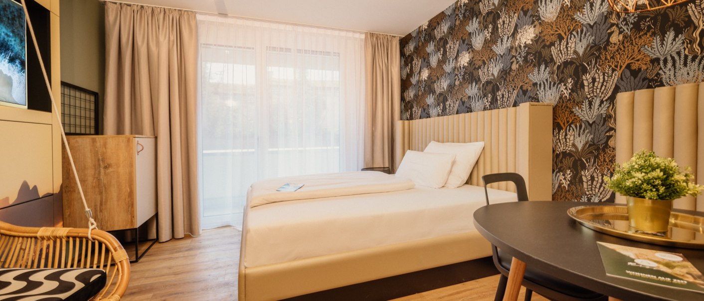 Modernes Apartmentzimmer mit Doppelbett, stilvoller Tapete, Hängesessel und Tisch mit Pflanze., © Rioca Stuttgart Posto 4 GmbH