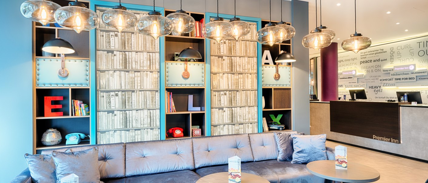 Moderne Lobby mit blauer Couch, runden Tischen und dekorativen Regalen. Hängelampen und ein Empfangstresen im Hintergrund., © Premier Inn Germany