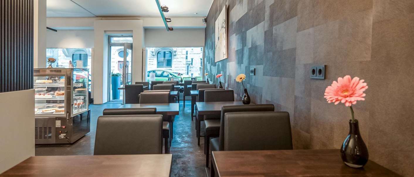 Modernes Café mit dunklen Tischen und Stühlen, Blumenvasen auf den Tischen und einer Kuchenvitrine im Hintergrund., © Café LIST, Stuttgart