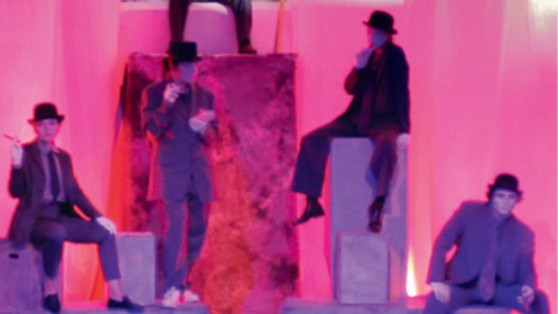 Fünf Personen in Anzügen und Hüten sitzen auf Podesten in rosa Licht. Die Szene wirkt theatralisch und mystisch., © Theaterschule an der Gmünder VHS