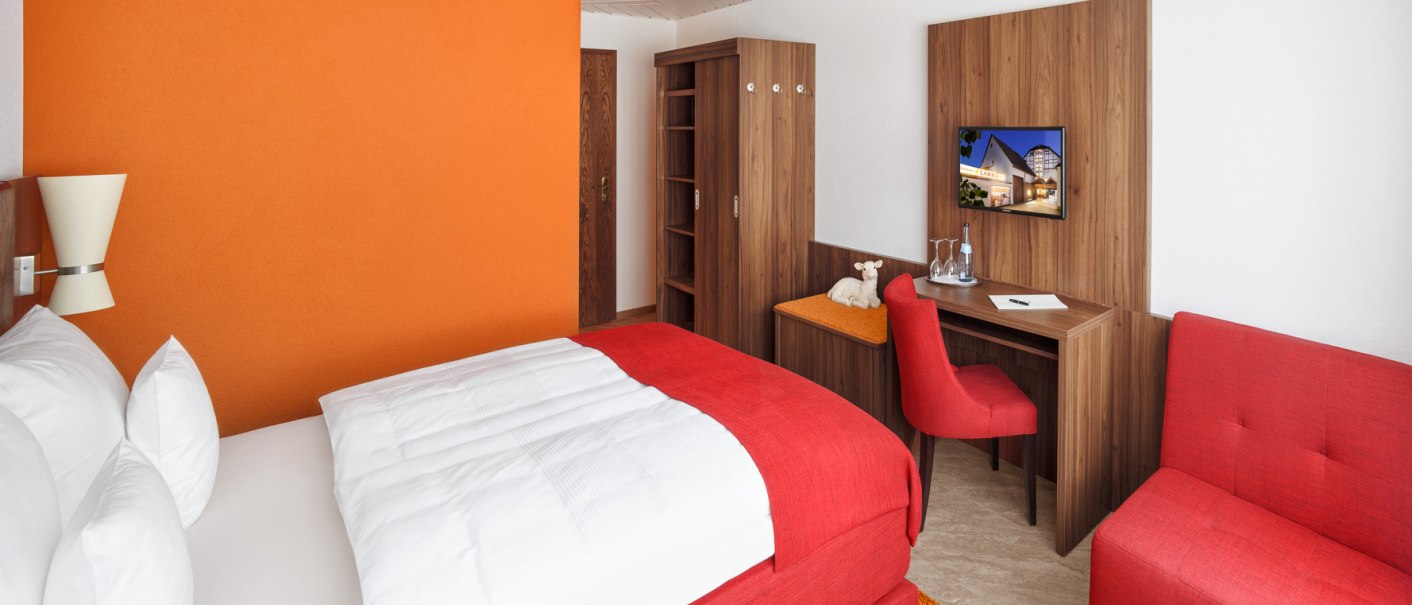 Modernes Hotelzimmer mit oranger Wand, rotem Bett und Sofa, Holzmöbeln, Schreibtisch und Fernseher., © Hotel Restaurant Lamm