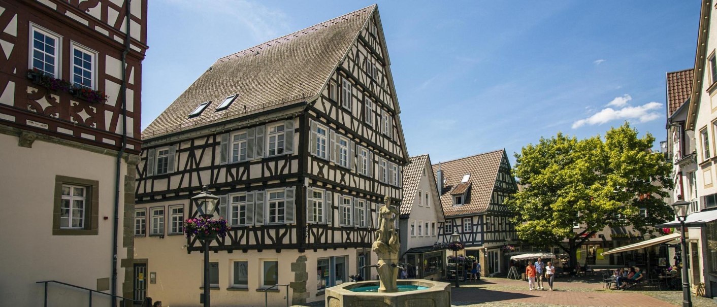 Fachwerkhäuser am Marktplatz von Backnang, mit einem Brunnen im Vordergrund und Bäumen im Hintergrund. Menschen spazieren und sitzen in Cafés., © Stuttgart-Marketing GmbH, Sarah Schmid Fachwerkhäuser am Marktplatz von Backnang, mit einem Brunnen im Vordergrund und Bäumen im Hintergrund. Menschen spazieren und sitzen in Cafés., © Stuttgart-Marketing GmbH, Sarah Schmid