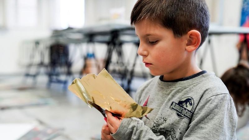 Ein Junge betrachtet konzentriert ein Stück goldene Folie in einem Raum mit Tischen im Hintergrund., © Kunstschule Labyrinth