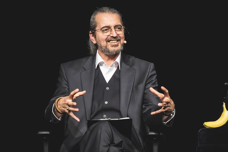 Ein Mann in einem Anzug sitzt auf einer B&uuml;hne, spricht gestikulierend in ein Mikrofon und l&auml;chelt. Neben ihm liegt eine Banane auf einem Tisch., &copy; Konzertkreis Triangel Compagnia Compostelli e.V.