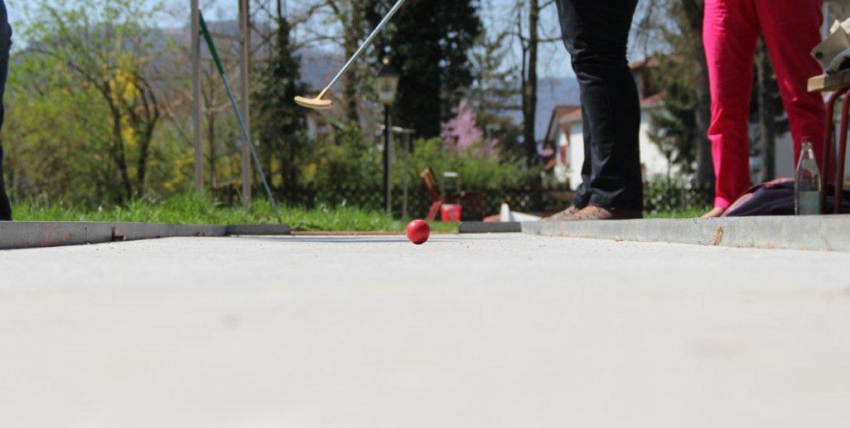 Zwei Personen spielen Minigolf auf einer Betonbahn im Freien. Eine rote Kugel rollt auf die Kamera zu. Im Hintergrund sind Bäume und ein Haus zu sehen.