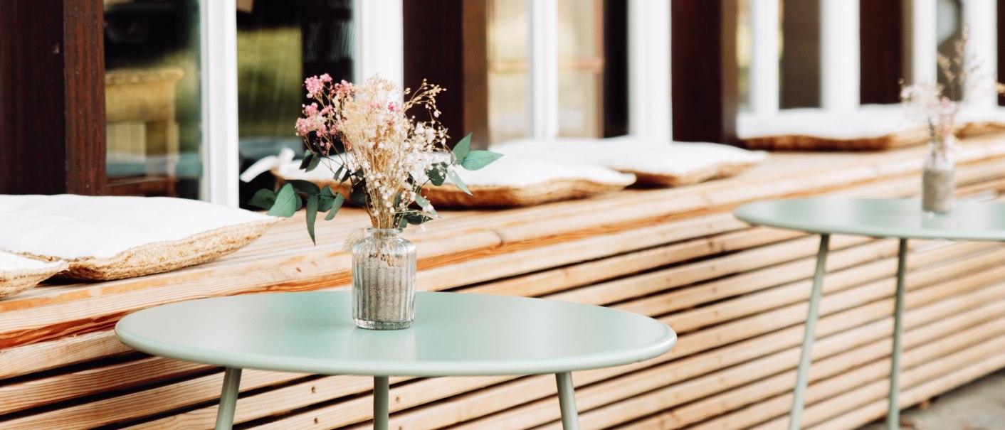 Zwei runde Tische mit Blumenvasen stehen vor einer Fensterfront im Außenbereich eines Cafés. Holzverkleidung und Sitzkissen sind sichtbar., © L. Prestle/ Documenting Love Zwei runde Tische mit Blumenvasen stehen vor einer Fensterfront im Außenbereich eines Cafés. Holzverkleidung und Sitzkissen sind sichtbar., © L. Prestle/ Documenting Love
