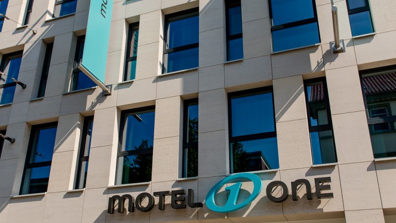 Moderne Fassade eines Gebäudes mit Motel One Logo und blauer Fahne, die im Wind weht. Klare Linien und große Fenster prägen das Design., © Motel One Stuttgart-Bad Cannstatt Moderne Fassade eines Gebäudes mit Motel One Logo und blauer Fahne, die im Wind weht. Klare Linien und große Fenster prägen das Design., © Motel One Stuttgart-Bad Cannstatt