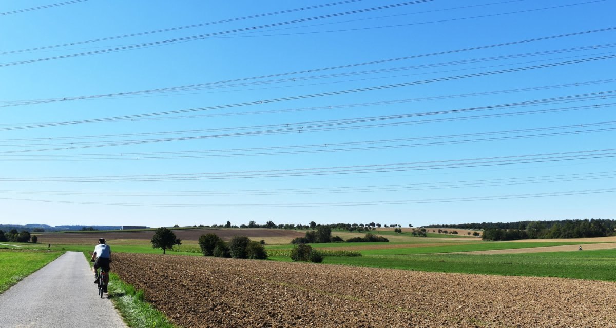 Ein Radfahrer fährt auf einer ländlichen Straße entlang eines Feldes. Der Himmel ist klar und blau, durchzogen von Stromleitungen., © Stadtmarketing Böblingen e.V. Ein Radfahrer fährt auf einer ländlichen Straße entlang eines Feldes. Der Himmel ist klar und blau, durchzogen von Stromleitungen., © Stadtmarketing Böblingen e.V.