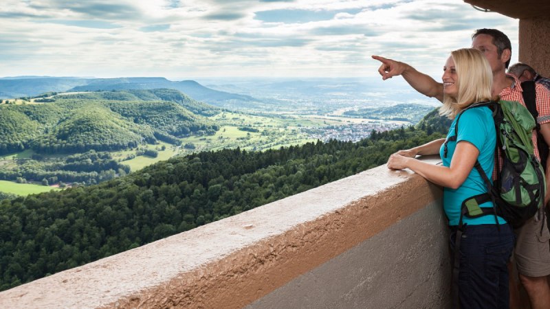 Zwei Personen stehen auf einem Aussichtsturm und blicken auf eine grüne Hügellandschaft. Eine Person zeigt in die Ferne., © hochgehberge