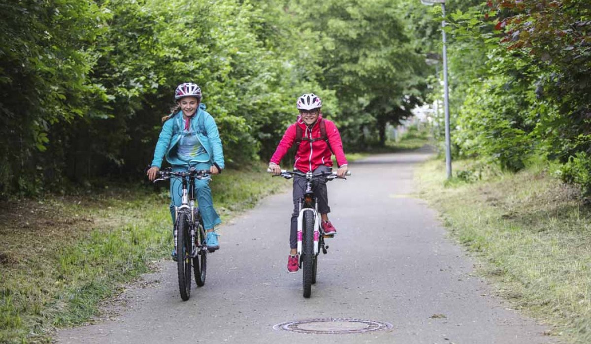 Zwei Kinder fahren auf einem Fahrradweg durch eine grüne Allee. Sie tragen Helme und bunte Kleidung. Der Weg ist von Bäumen gesäumt., © Schwäbische Alb Tourismusverband e.V.