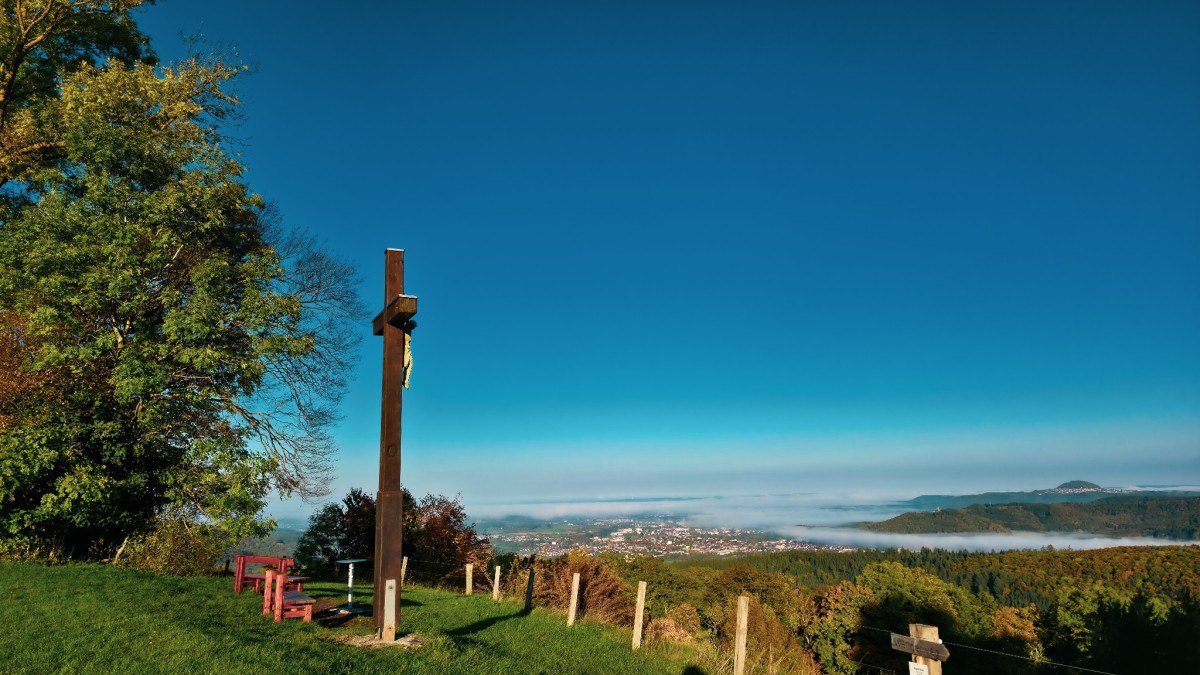 Ein Holzkreuz steht auf einer gr&uuml;nen Wiese mit Blick auf ein nebelverhangenes Tal und eine Stadt in der Ferne. Der Himmel ist klar und blau., &copy; Distretto di G&ouml;ppingen