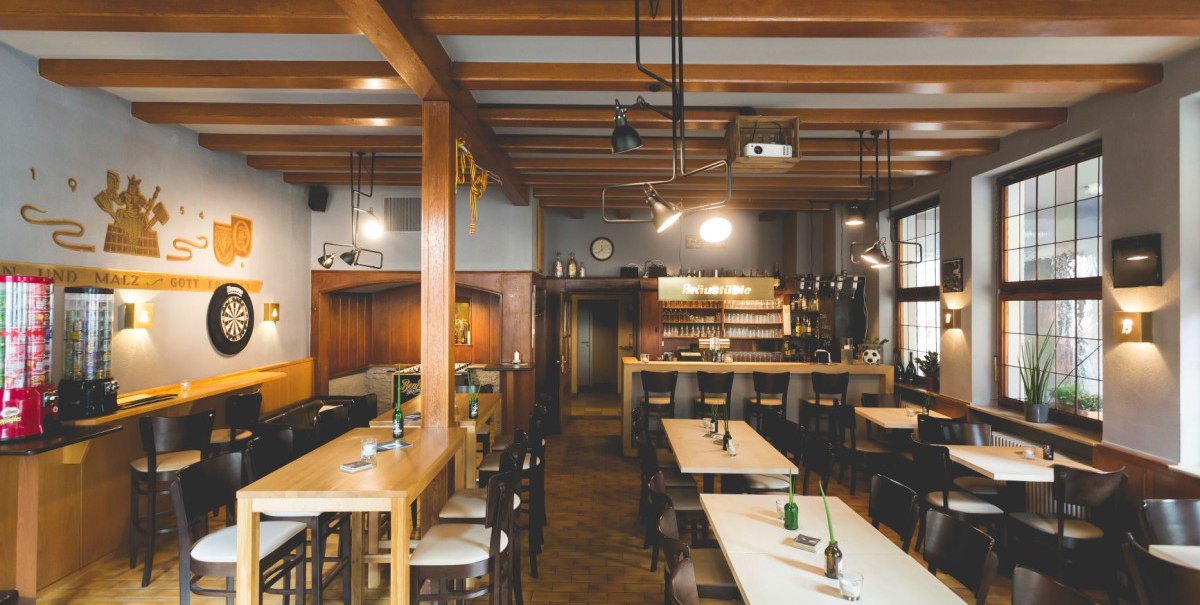 Gemütliche Bar mit Holzmöbeln, Barhockern, Dartboard und dekorativen Elementen an der Wand. Große Fenster lassen Tageslicht herein.