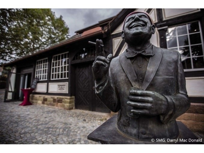 Bronzestatue eines lachenden Mannes mit Fliege vor einem historischen Fachwerkgeb&auml;ude. Kopfsteinpflaster im Vordergrund, B&auml;ume im Hintergrund., &copy; Stuttgart Marketing GmbH