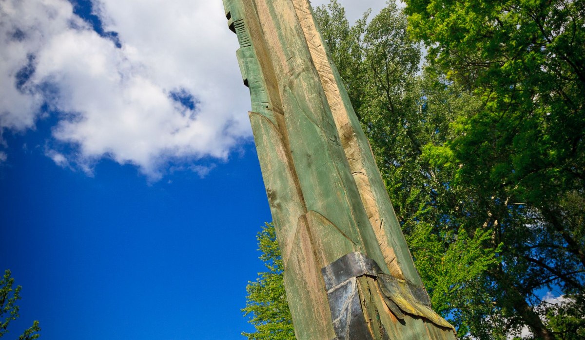 Eine hohe Baumskulptur aus Holz ragt in den blauen Himmel, umgeben von grünen Bäumen und Häusern im Hintergrund., © Remstal Tourismus e.V.