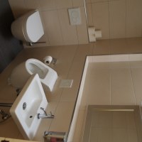 Ein modernes Badezimmer mit Toilette, Bidet und Waschbecken, alles in Weiß gehalten. Die Wände sind mit beigen Fliesen verkleidet., © TOMAS Ein modernes Badezimmer mit Toilette, Bidet und Waschbecken, alles in Weiß gehalten. Die Wände sind mit beigen Fliesen verkleidet., © TOMAS
